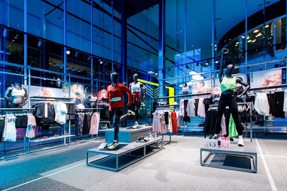 A spacious, modern clothing store with mannequins displayed on raised platforms. The mannequins are dressed in athletic wear, including leggings, sports bras, and jackets. Large glass windows provide an outside view, letting in natural light. Clothing racks filled with various apparel items, including leggings, shorts, and tops, line the walls. The overall setting is clean and organized.