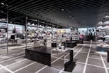 A modern sneaker store with sleek black shelving displaying various athletic shoes and sports apparel. Central seating areas and spotlights provide a contemporary retail ambiance. The signage prominently features a famous sportswear slogan in the background.