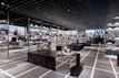 A modern sneaker store with sleek black shelving displaying various athletic shoes and sports apparel. Central seating areas and spotlights provide a contemporary retail ambiance. The signage prominently features a famous sportswear slogan in the background.