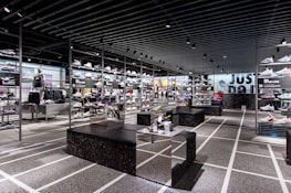 A modern sneaker store with sleek black shelving displaying various athletic shoes and sports apparel. Central seating areas and spotlights provide a contemporary retail ambiance. The signage prominently features a famous sportswear slogan in the background.