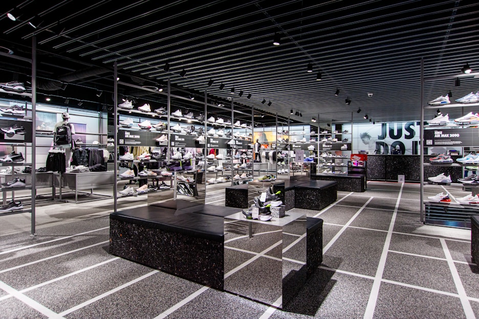 A modern sneaker store with sleek black shelving displaying various athletic shoes and sports apparel. Central seating areas and spotlights provide a contemporary retail ambiance. The signage prominently features a famous sportswear slogan in the background.