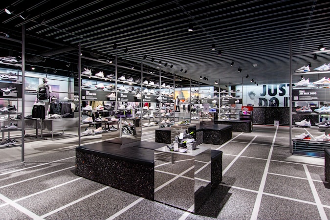 A modern sneaker store with sleek black shelving displaying various athletic shoes and sports apparel. Central seating areas and spotlights provide a contemporary retail ambiance. The signage prominently features a famous sportswear slogan in the background.