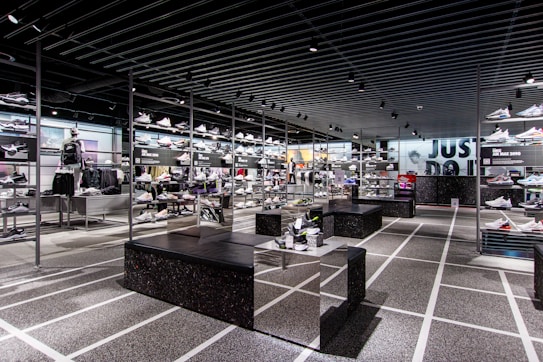 A modern sneaker store with sleek black shelving displaying various athletic shoes and sports apparel. Central seating areas and spotlights provide a contemporary retail ambiance. The signage prominently features a famous sportswear slogan in the background.