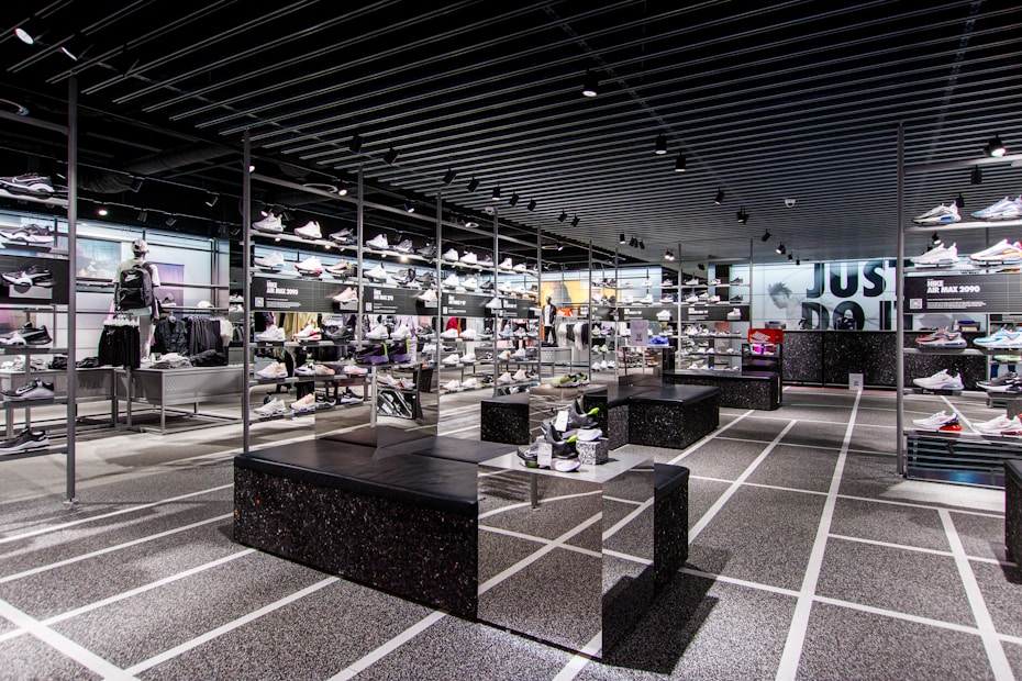 A modern sneaker store with sleek black shelving displaying various athletic shoes and sports apparel. Central seating areas and spotlights provide a contemporary retail ambiance. The signage prominently features a famous sportswear slogan in the background.