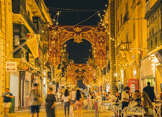 A vibrant nighttime street scene with people dining outdoors. Brightly lit decorations and ornate banners hang between tall, historic buildings. Tables are set with people enjoying meals, and ambient string lights illuminate the area. There is a bustling, lively atmosphere with people walking by and engaging in conversation.