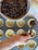 A hand is scooping a mixture of chopped nuts and dried fruits from a bowl into pastry shells in a muffin tray. The mixture appears to be rich and dark, likely intended for baking.