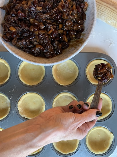 A hand is scooping a mixture of chopped nuts and dried fruits from a bowl into pastry shells in a muffin tray. The mixture appears to be rich and dark, likely intended for baking.