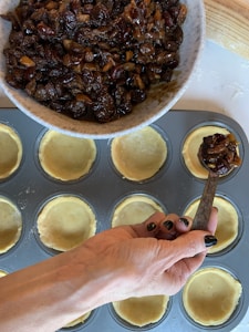 A hand is scooping a mixture of chopped nuts and dried fruits from a bowl into pastry shells in a muffin tray. The mixture appears to be rich and dark, likely intended for baking.