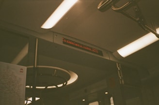 The interior of a subway train with fluorescent lights and a digital sign displaying text. A subway map is partially visible on the left side. Metal bars and handles are present, indicating it is a commuter environment.