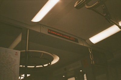 The interior of a subway train with fluorescent lights and a digital sign displaying text. A subway map is partially visible on the left side. Metal bars and handles are present, indicating it is a commuter environment.