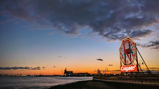 A large clock with a prominent 'Colgate' logo sits at the edge of a waterfront, with a colorful sunset painting the sky in hues of orange, blue, and purple. The silhouette of industrial buildings and a railing along a walkway are visible.