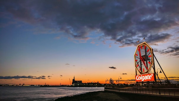 A large clock with a prominent 'Colgate' logo sits at the edge of a waterfront, with a colorful sunset painting the sky in hues of orange, blue, and purple. The silhouette of industrial buildings and a railing along a walkway are visible.