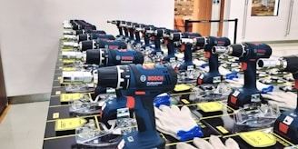Multiple Bosch professional power drills are neatly arranged in rows on a display table. Each drill is supported by a clear plastic stand, and there are white gloves placed nearby. The setting appears to be indoors, possibly a store or showroom, with an orange chair and glass door visible in the background.