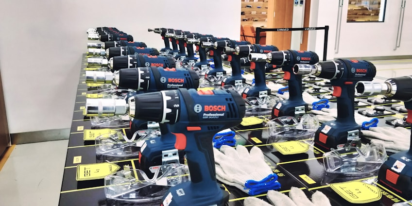 Multiple Bosch professional power drills are neatly arranged in rows on a display table. Each drill is supported by a clear plastic stand, and there are white gloves placed nearby. The setting appears to be indoors, possibly a store or showroom, with an orange chair and glass door visible in the background.