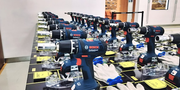 Multiple Bosch professional power drills are neatly arranged in rows on a display table. Each drill is supported by a clear plastic stand, and there are white gloves placed nearby. The setting appears to be indoors, possibly a store or showroom, with an orange chair and glass door visible in the background.