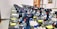 Multiple Bosch professional power drills are neatly arranged in rows on a display table. Each drill is supported by a clear plastic stand, and there are white gloves placed nearby. The setting appears to be indoors, possibly a store or showroom, with an orange chair and glass door visible in the background.