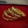 A set of intricately designed gold bangles with detailed patterns is placed on a plush red surface. The bangles exhibit a polished, reflective finish, enhancing their luxurious appearance.