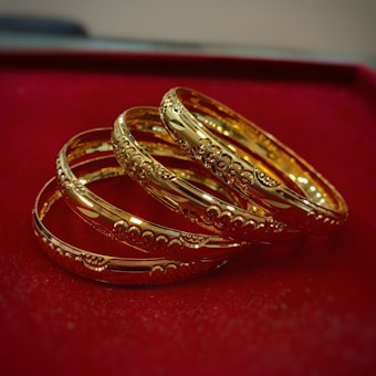 A set of intricately designed gold bangles with detailed patterns is placed on a plush red surface. The bangles exhibit a polished, reflective finish, enhancing their luxurious appearance.