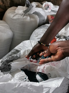 A pair of hands holding small blue beads over an open bag, surrounded by large white sacks in a dimly lit setting. The hands are adorned with colorful beaded bracelets.