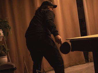 A person wearing a dark outfit and cap is playing table tennis. The room is dimly lit with beige curtains as the background. There's a potted plant visible on the left side and a table tennis table with a paddle in the right hand of the person.