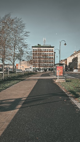 A wide pedestrian and bike path flanked by grass and trees runs towards a multi-story building in an urban area. The building has the word 'VERITAS' on top and is surrounded by other smaller structures. The sky is clear with a faint moon visible. Streetlamps line the path, and a brightly colored advertisement is displayed on a bus stop.