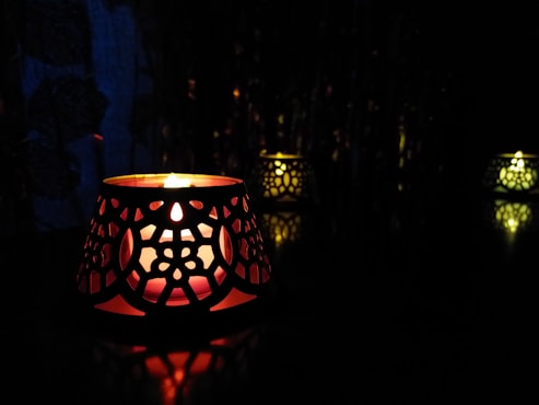 A candle inside an intricately designed holder casts a warm, soft glow, creating reflections on a dark surface. The ambiance is cozy and intimate, with additional similar candle holders faintly visible in the background.
