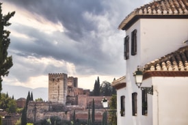 A historic fortress is seen in the background, featuring large towers and ancient stone walls, surrounded by lush greenery and tall cypress trees. In the foreground, a traditional building with a tiled roof and whitewashed walls is visible, contributing to the architectural charm of the scene. The sky is dramatic, filled with dark, heavy clouds that suggest an impending storm.