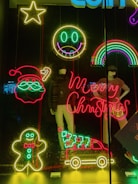 A vibrant display of neon lights featuring Christmas-themed designs, including a Santa face, a gingerbread man, and a vehicle carrying a tree. Mannequins in the background are dressed in winter apparel. A cheerful smiley face and a rainbow add to the festive mood.