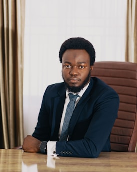 A man in a suit sits at a wooden desk, looking directly at the camera. He is wearing a dark navy suit, a white shirt, and a blue tie. His arms are resting on the desk and he wears a watch on his left wrist. Behind him, there is an office chair and light-colored curtains with soft natural light filtering through.