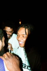 A group of young men is enjoying a moment together at night. They are smiling and appear to be engaged in lighthearted conversation. The lighting highlights their faces while the background remains dark, emphasizing their expressions and interactions.