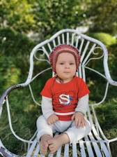 A baby sits on a white wrought iron chair in a garden setting, wearing a red shirt with an 'S' logo and white polka-dotted pants. The baby has a maroon bonnet on and the background is lush with green foliage.