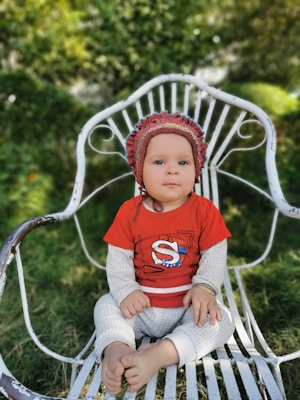 A baby sits on a white wrought iron chair in a garden setting, wearing a red shirt with an 'S' logo and white polka-dotted pants. The baby has a maroon bonnet on and the background is lush with green foliage.