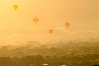 Hot air balloons are floating above a lush green landscape, with the warm glow of the sun casting a golden hue over the scene. The horizon is hazy, adding a dreamy, ethereal quality to the image.