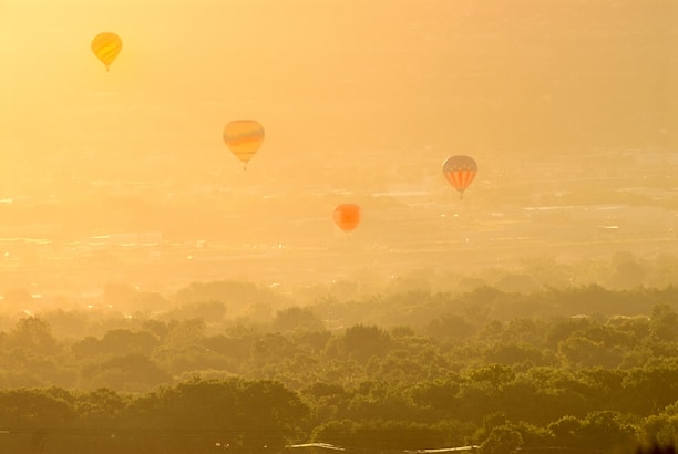 Hot air balloons are floating above a lush green landscape, with the warm glow of the sun casting a golden hue over the scene. The horizon is hazy, adding a dreamy, ethereal quality to the image.
