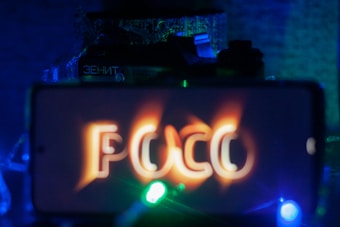 A smartphone screen displaying the text 'POCO' with a glowing, fiery effect. The background includes a vintage camera partially visible with the words 'ZENIT'. The scene is illuminated with blue, green, and orange lighting, creating a mysterious and vibrant atmosphere.