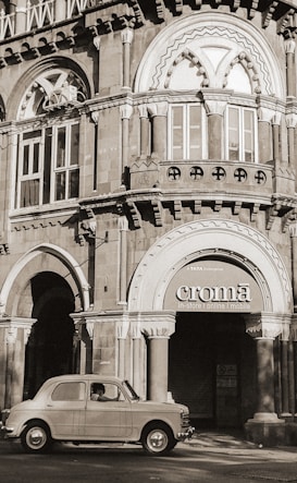 A vintage car is parked in front of a historic building with elaborate architecture. The building features arched windows, intricate stonework, and a sign for a store named 'Croma'. The image has a sepia tone, giving it a nostalgic feel.