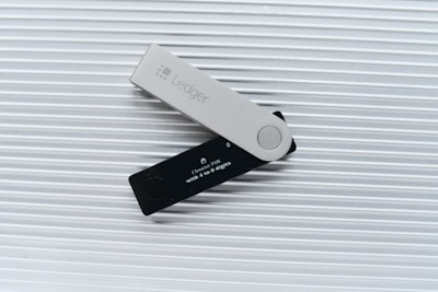 A Ledger hardware wallet is positioned on a ridged light gray surface. It features a contrasting design with a black part displaying a small screen and a gray cover imprinted with the Ledger logo. The screen contains text about choosing a PIN.