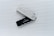 A Ledger hardware wallet is positioned on a ridged light gray surface. It features a contrasting design with a black part displaying a small screen and a gray cover imprinted with the Ledger logo. The screen contains text about choosing a PIN.