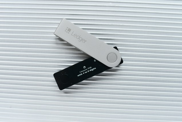 A Ledger hardware wallet is positioned on a ridged light gray surface. It features a contrasting design with a black part displaying a small screen and a gray cover imprinted with the Ledger logo. The screen contains text about choosing a PIN.