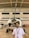 A young man stands in front of a small propeller aircraft inside a hangar. The hangar has high ceilings with exposed beams and several windows. The man is wearing a white shirt and a lanyard with an ID card. The aircraft has a shiny propeller and is predominantly white with red accents.