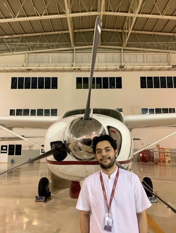 A young man stands in front of a small propeller aircraft inside a hangar. The hangar has high ceilings with exposed beams and several windows. The man is wearing a white shirt and a lanyard with an ID card. The aircraft has a shiny propeller and is predominantly white with red accents.