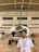 A young man stands in front of a small propeller aircraft inside a hangar. The hangar has high ceilings with exposed beams and several windows. The man is wearing a white shirt and a lanyard with an ID card. The aircraft has a shiny propeller and is predominantly white with red accents.