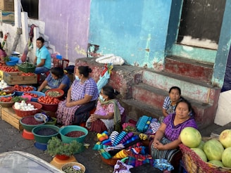 A group of women and a man are situated on a street corner, surrounded by baskets and crates filled with fresh produce, such as tomatoes, onions, beans, and herbs. They are sitting on the pavement with colorful woven textiles on display for sale. The women wear traditional patterned clothing, and one of them is wearing a mask. The background features worn-out blue and purple walls, with a set of steps leading to a closed door.