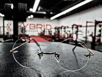 A pair of round metal-framed glasses is placed on a black surface in a gym setting. The background displays blurred gym equipment and weights with a red carpet, and the word 'HYBRID' is visible on the wall.