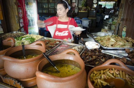A woman wearing a red apron is serving food in a restaurant setting. Various dishes are displayed, including curry in clay pots, a platter of vegetables, and trays containing different meats and eggs. The environment appears to be a casual, local eatery with a rustic wooden interior. There are metal trays, ladles, and utensils, and the woman is in motion, suggesting she is busy serving or preparing dishes.