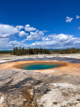 A large geothermal hot spring is featured, with vibrant water that transitions from deep blue in the center to green and then bright orange at the edges. The spring is surrounded by a barren, rocky landscape. In the background, there is a line of dense evergreen trees under a bright blue sky filled with some fluffy white clouds.
