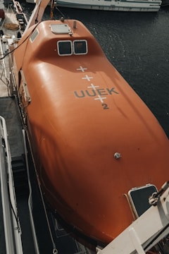 A bright orange lifeboat is securely mounted on a ship. The lifeboat features various mechanical components and is marked with identification codes. It is positioned next to the dark, reflective water of a harbor, with parts of another boat visible in the background.