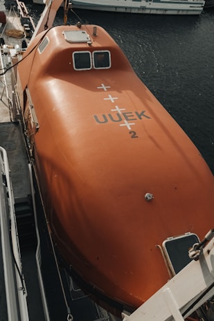 A bright orange lifeboat is securely mounted on a ship. The lifeboat features various mechanical components and is marked with identification codes. It is positioned next to the dark, reflective water of a harbor, with parts of another boat visible in the background.