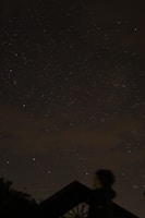 A night sky filled with numerous stars, with a dimly lit foreground showing a blurry silhouette of a person and the outline of a structure. The scene conveys a quiet and serene atmosphere, with the vastness of the universe apparent overhead.