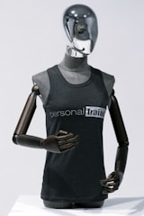A metallic mannequin with a smooth, reflective head and dark, articulated arms is wearing a sleeveless shirt that reads 'personal trainer'. The mannequin stands upright against a plain light background, emphasizing the sleek design.